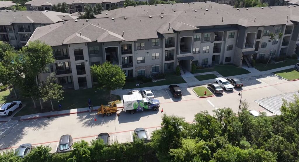 view of apartment building from the sky with cars parked in front with a blue, green and white chipper truck and yellow chipper behind it; Tree Trimming, Tree removal